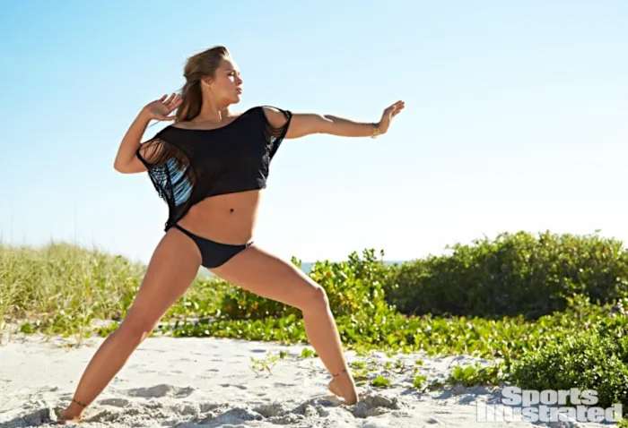 Ronda Rousey performs a martial arts pose in the sand wearing a black sheer shirt and black bikini bottoms.